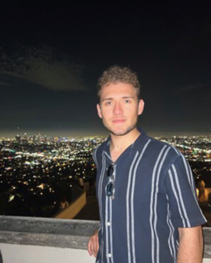 A man in a stiped shirt posing in front of a well lit cityscape at night.