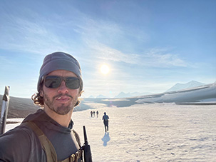 A selfie-style picture of a man in sunglasses and a winter hat with a snowscape and mountains behind him. He has a handheld radio attached to the harness on his chest. Other people are in the background using trekking poles. The sun is shining brightly.