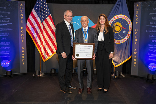 Image of Tananbaum on a stage with NASA backdrop. He is standing in between a man and a woman while holding an award certificate and wearing a gold medal on a blue ribbon around his neck.