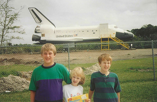 A slightly grainy with age photo of three children posing in front of the length of Space Shuttle Explorer. The childen are squinting towards the camera on an obviously bright day.