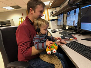 John is smiling, with his son on his lap at a computer terminal. The child staring intently at the keyboard while wearing headphones and holding a stuffed toy car in his hand.